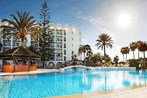 Kuva lomakeskuksesta, jossa on ulkouima-allas, puusilta altaan yli, palmuja ja valkoinen rakennus taustalla. (Tekoälyn luoma teksti)