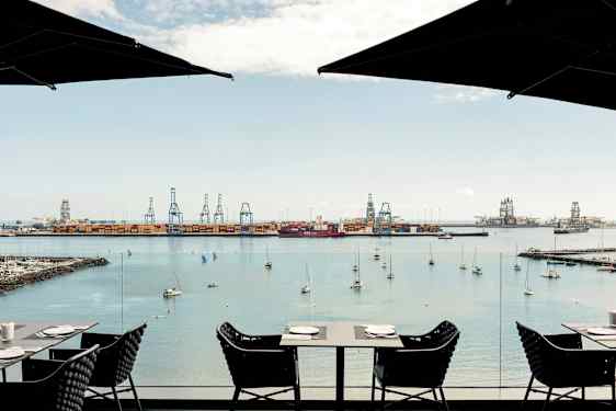 Näkymä satamaan, jossa on nostureita ja kiinnittyneitä veneitä. Etualalla on pöytiä ja tuoleja kahvilasta. (Tekoälyn luoma teksti)