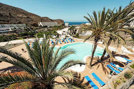 Näkymä hotellin uima-altaalle, jota ympäröivät aurinkotuolit ja palmupuut, taustalla vuoret ja meri. (Tekoälyn luoma teksti)