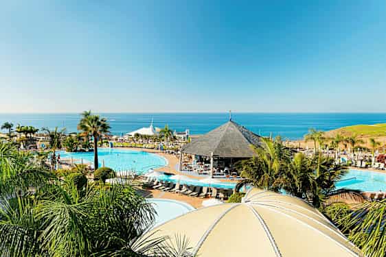 Lomakohde rannikolla, jossa on uima-altaita, paviljonki ja palmuja sekä meri taustalla. (Tekoälyn luoma teksti)