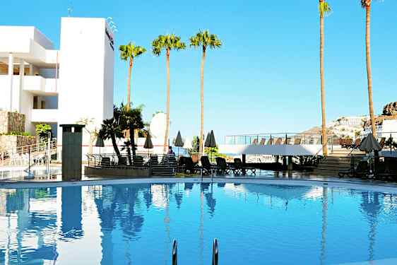 Uima-allas, jota ympäröivät aurinkotuolit, aurinkovarjot ja palmupuut, taustalla rakennus ja kirkas sininen taivas. (Tekoälyn luoma teksti)