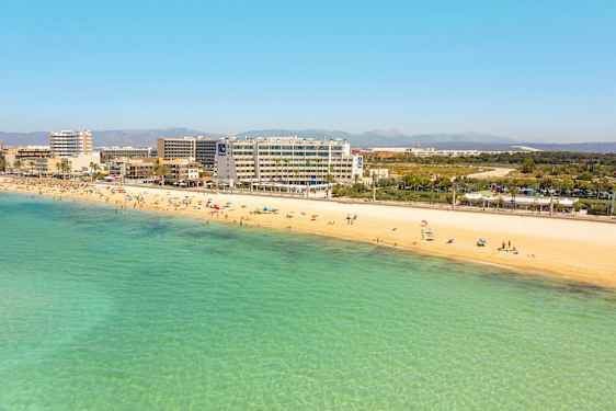 Hiekkaranta, jossa uimareita ja kirkasta turkoosia vettä. Hotelleja ja rakennuksia taustalla. (Tekoälyn luoma teksti)