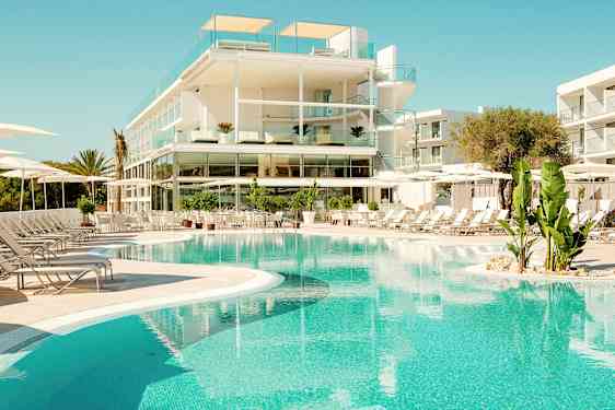 Moderni hotelli, jossa on lasiparvekkeet, sijaitsee kirkkaan sinisen uima-altaan vieressä, jota ympäröivät aurinkotuolit ja vihreät kasvit. (Tekoälyn luoma teksti)