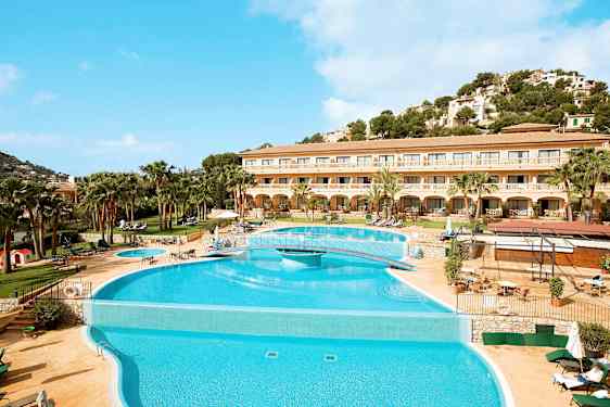 Kuvassa on hotelli, jonka edessä on suuri uima-allas ja ympärillä terassi aurinkotuoleineen. Taustalla on vehreä mäki ja kirkkaan sininen taivas. (Tekoälyn luoma teksti)