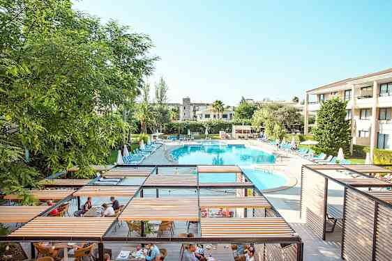 Lomakeskus, jossa on suuri soikea uima-allas, jota ympäröivät aurinkotuolit ja aurinkovarjot, sekä ruokailualue pergoloilla. (Tekoälyn luoma teksti)