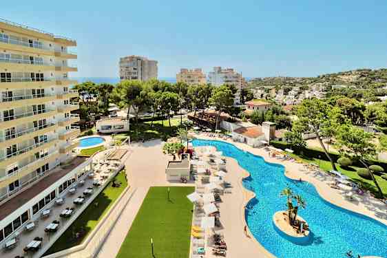 Hotellirakennus uima-altaan vieressä, jossa on aurinkotuoleja ja varjoja, sekä ympäröivä kasvillisuus ja näkymä kaupunkiin. (Tekoälyn luoma teksti)