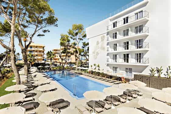 Kuva hotellialueesta, jossa on suuri uima-allas ympäröitynä aurinkotuoleilla ja varjoilla. Valkoinen rakennus, jossa on useita parvekkeita, sekä puita taustalla. (Tekoälyn luoma teksti)