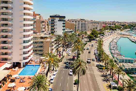 Kuva, jossa näkyy katu palmujen kera, uima-allas rakennuskompleksin luona ja satama veneineen. (Tekoälyn luoma teksti)