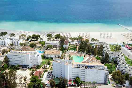 Ilmakuva hotellialueesta lähellä rantaa, jossa hiekan päällä on aurinkovarjoja ja kirkkaan sininen merimaisema. (Tekoälyn luoma teksti)