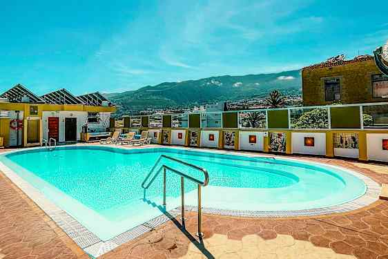 Ulkoallas aurinkotuoleilla aurinkoisessa ympäristössä, jota ympäröivät rakennukset ja taustalla vuoret. (Tekoälyn luoma teksti)