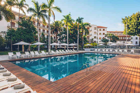 Ulkouima-allas, jota ympäröivät aurinkotuolit, varjot ja vehreät puut, taustalla rakennus. (Tekoälyn luoma teksti)