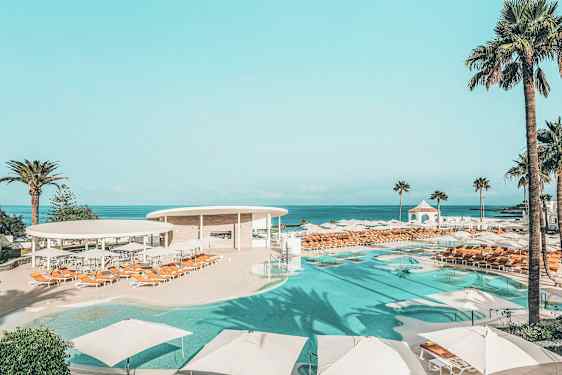 Uima-allasalue, jossa on aurinkovarjoja ja aurinkotuoleja, ympäröitynä palmupuilla ja merinäköalalla. (Tekoälyn luoma teksti)