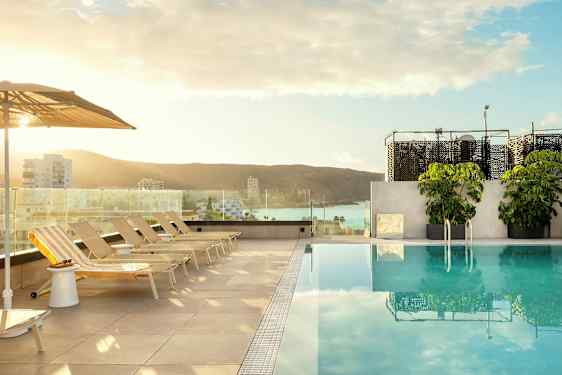 Näkymä uima-altaalle, jossa on aurinkotuoleja ja aurinkovarjoja; taustalla näkyy kaupunki ja meri. (Tekoälyn luoma teksti)