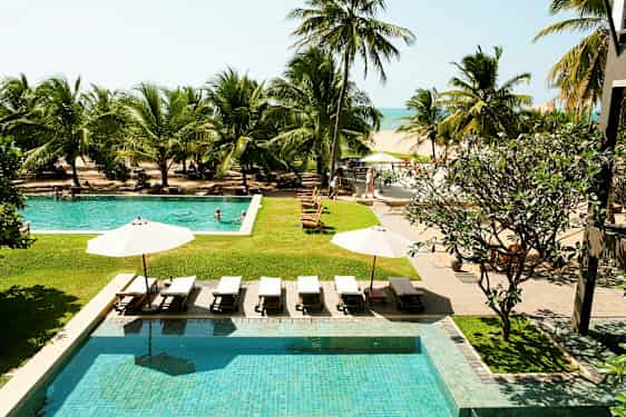 Kuvassa on hotellin uima-allasalue aurinkotuoleineen ja aurinkovarjoineen, trooppisten palmujen ympäröimänä, näkymällä rannalle ja merelle. (Tekoälyn luoma teksti)