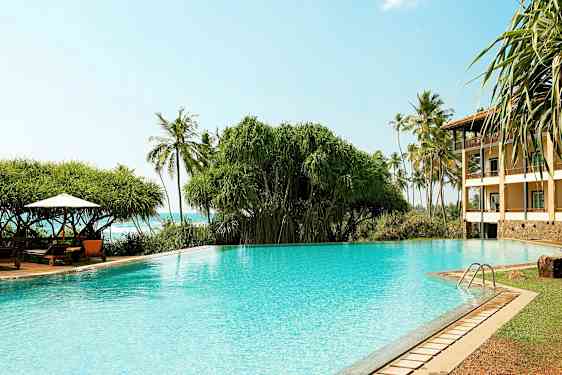 Kirkas sininen uima-allas kasvillisuuden ja palmujen taustalla sekä rakennus oikealla. (Tekoälyn luoma teksti)