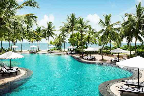 Kuva uima-altaasta, jota ympäröivät palmut ja aurinkotuolit sateenvarjoineen lähellä merta. (Tekoälyn luoma teksti)