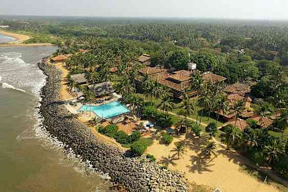 Näkymä rannikkoviivalle, jossa hotellialue, johon kuuluu uima-allas, tiilikattoisia rakennuksia sekä vehreitä ja kookospuiden ympäröimiä maisemia. (Tekoälyn luoma teksti)