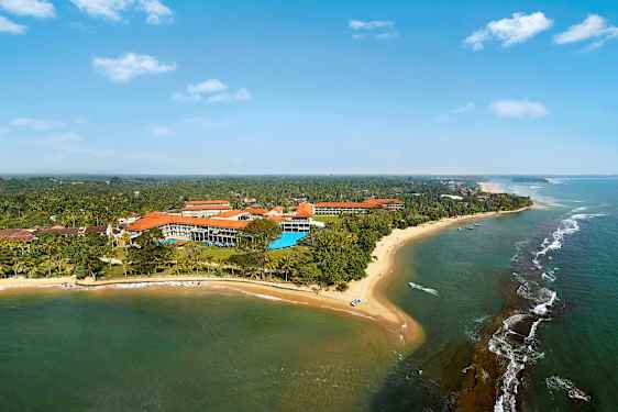 Ilmakuva rantaviivasta, jossa on hiekkaranta, meri ja rakennuksia vehreyden keskellä. (Tekoälyn luoma teksti)