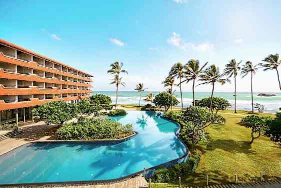 Hotellirakennus rannalla, jossa on suuri uima-allas, palmuja ja meri taustalla. (Tekoälyn luoma teksti)