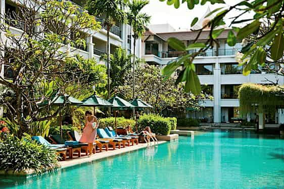 Kuva ulkouima-altaasta, jonka ympärillä on aurinkotuoleja, aurinkovarjoja ja kasveja, taustalla rakennus parvekkeineen. (Tekoälyn luoma teksti)