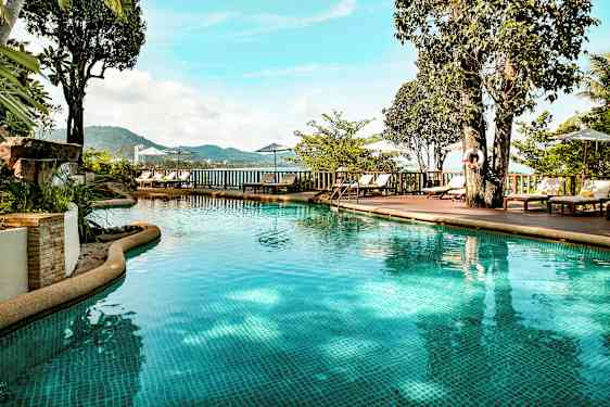 Ulkoallas kirkkaalla vedellä, jota ympäröivät aurinkotuolit, aurinkovarjot ja vehreys. Näköala paljastaa vuoria ja merta taustalla. (Tekoälyn luoma teksti)