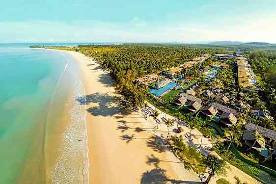 Ilmakuva pitkästä hiekkarannasta turkoosin veden äärellä ja lomakeskuksesta, jossa on bungaloweja ja uima-altaita. (Tekoälyn luoma teksti)