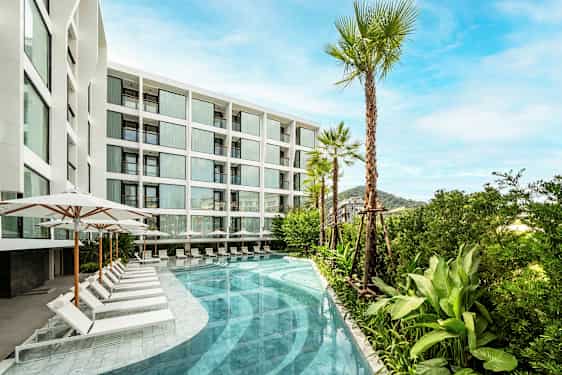 Kuva modernista valkoisesta rakennuksesta, jossa on suuret ikkunat. Rakennuksen edustalla on uima-allas aurinkotuoleineen ja varjoineen sekä palmuja ja vehreyttä. (Tekoälyn luoma teksti)