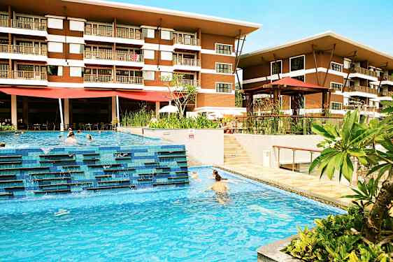 Hotelli, jossa on kaksi kerrosta, näkyy ulkouima-altaan takana, jossa ihmiset uivat. Allasalueen ympärillä on vihreitä kasveja ja terassialueita. (Tekoälyn luoma teksti)