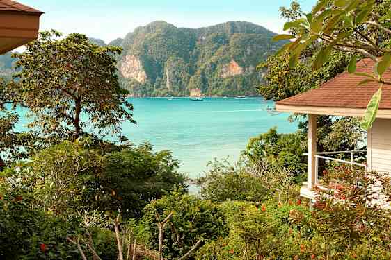 Näkymä lahdelle, jossa on kirkasta sinistä vettä, ympäröitynä vehreällä kasvillisuudella ja vuorilla, oikealla näkyy osa rakennusta. (Tekoälyn luoma teksti)