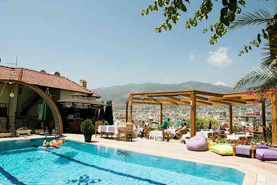 Ulkouima-allas hotellilla, jossa aurinkotuoleja ja aurinkovarjoja. Puinen pergola ja näkymä kaupunkiin ja vuorille taustalla. (Tekoälyn luoma teksti)