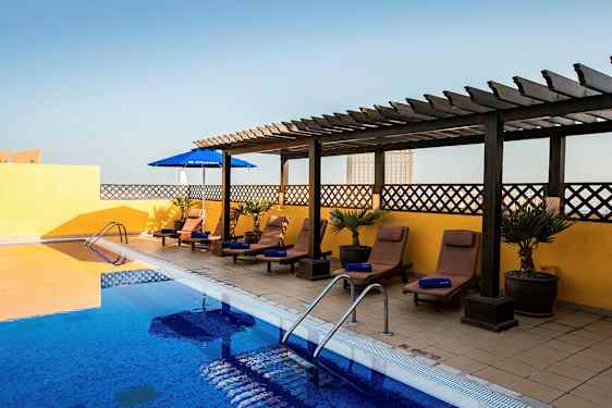 Ulkouima-allas, jossa sinistä vettä, ympärillä aurinkotuoleja ja aurinkovarjoja. Aurinkotuolit ovat laatoitetulla lattialla, jonka taustalla keltainen seinä ja puinen pergola. (Tekoälyn luoma teksti)