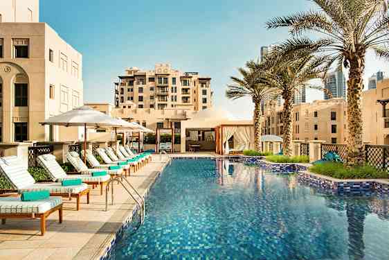 Ulkouima-allas, jota ympäröivät aurinkotuolit, aurinkovarjot ja palmupuut rakennusten vieressä. (Tekoälyn luoma teksti)