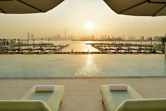 Näkymä infinity-altaalta pienvenesatamaan ja kaupungin siluettiin auringonlaskun aikaan, etualalla kaksi aurinkovuodetta. (Tekoälyn luoma teksti)