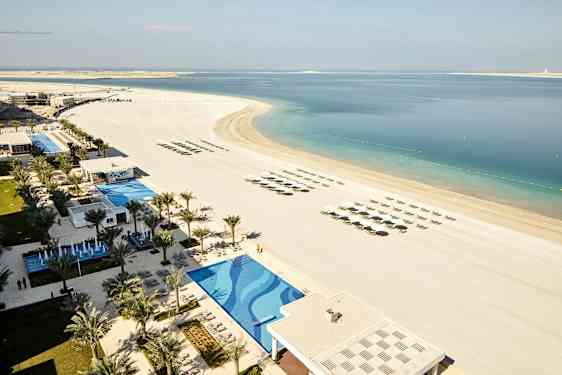 Kuvassa on ranta, jossa on aurinkovarjoja ja aurinkotuoleja riveissä, uima-altaan ja lomakeskusalueen vieressä palmuineen. (Tekoälyn luoma teksti)