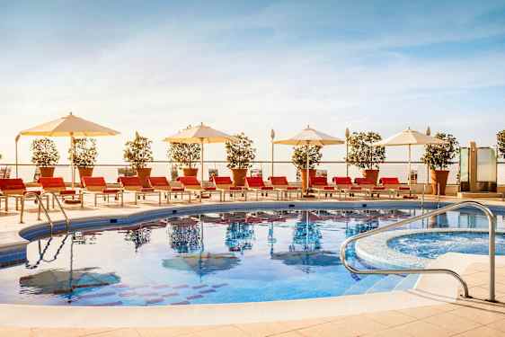 Ulkouima-allas, jonka ympärillä on punaisia aurinkotuoleja, valkoisia aurinkovarjoja ja ruukkukasveja. (Tekoälyn luoma teksti)