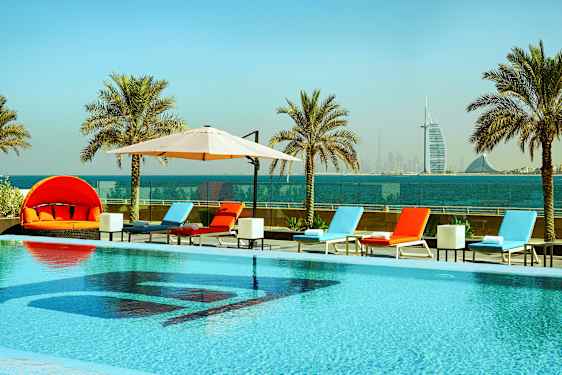 Uima-allas aurinkotuoleineen ja aurinkovarjoineen, ympärillä palmuja, taustalla merinäkymä ja kaupungin siluetti. (Tekoälyn luoma teksti)