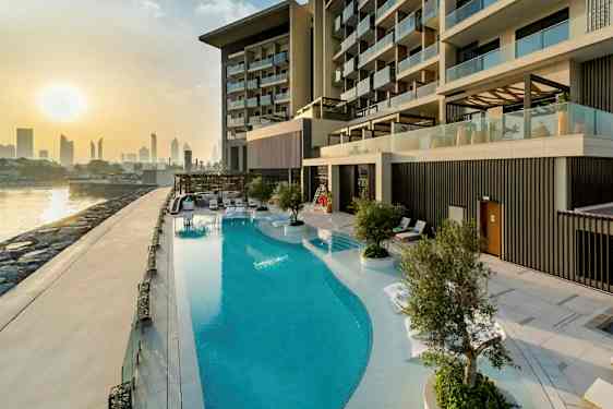 Kuva ylellisestä hotelliuima-altaasta rakennuksen vieressä, jossa on moderneja parvekkeita. Taustalla näkyy kaupunkisiluetti ja auringonlasku. (Tekoälyn luoma teksti)