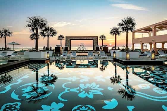 Kuva uima-altaasta, jossa on koristeellinen kuvio. Ympärillä aurinkotuoleja, palmuja ja taustalla auringonlasku. (Tekoälyn luoma teksti)