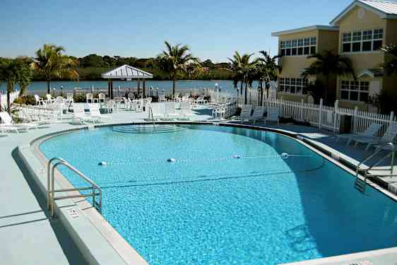 Kuva ulkouima-altaasta, jota ympäröivät aurinkotuolit ja päivänvarjot. Taustalla näkyy pieni järvi viheralueiden ympäröimänä. (Tekoälyn luoma teksti)