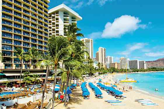 Hiekkaranta, jolla on aurinkotuoleja ja sinisiä aurinkovarjoja, ympärillä korkeita hotellirakennuksia ja palmuja. (Tekoälyn luoma teksti)