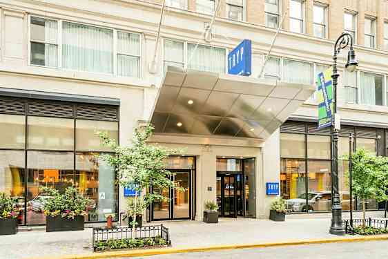 Hotellin sisäänkäynti, jossa on kyltti "TRYP". Rakennuksessa on suuret ikkunat ja istutuksia. (Tekoälyn luoma teksti)