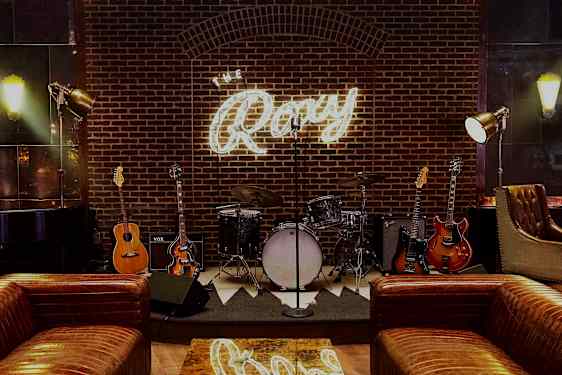 Lava, jossa on soittimia, kuten rummut ja kitaroita, tiiliseinän edessä, jossa on valaistu kyltti "The Roxy". (Tekoälyn luoma teksti)