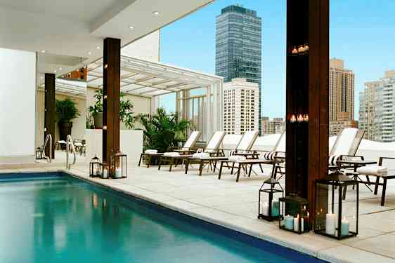 Moderni ulkouima-allas, jossa aurinkotuoleja ja sytytettyjä lyhtyjä kaupunkiympäristössä. (Tekoälyn luoma teksti)