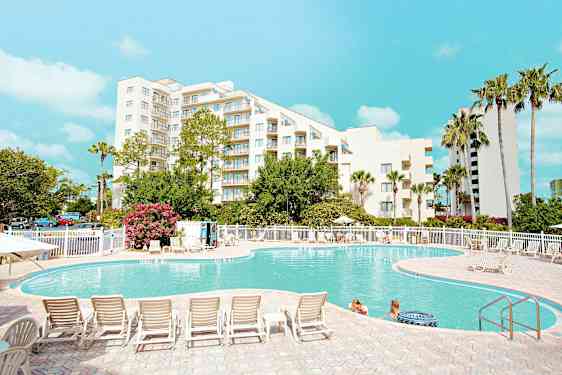 Ulkouima-allas valkoisen rakennuksen edustalla, ympärillä puita ja aurinkotuoleja, ihmisiä vedessä ja kirkas sininen taivas yläpuolella. (Tekoälyn luoma teksti)