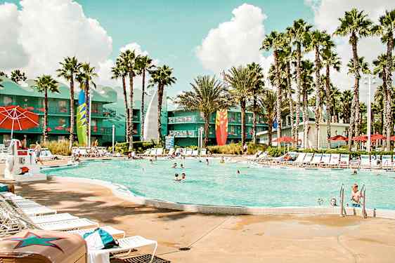 Ulkouima-allas, jonka ympärillä on aurinkotuoleja, aurinkovarjoja ja palmuja. Taustalla näkyy värikkäisiin yksityiskohtiin koristeltu rakennus. (Tekoälyn luoma teksti)