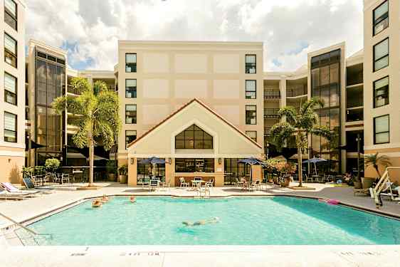 Kuva hotellista, jonka etualalla on uima-allas. Ihmiset uivat ja rentoutuvat allasalueella. Hotellin julkisivu näkyy taustalla ja ympärillä on palmuja. (Tekoälyn luoma teksti)