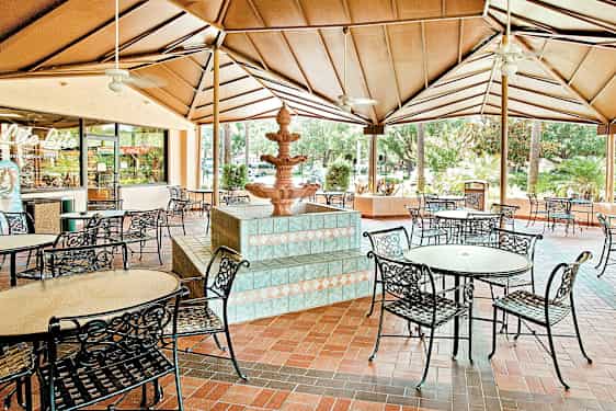 Katettu terassi, jossa on pyöreitä pöytiä ja metallituoleja sekä suihkulähde keskellä. (Tekoälyn luoma teksti)