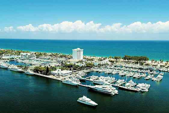 Satama, jossa veneitä ja jahteja rannikon äärellä, takana rakennus ja hotelli. (Tekoälyn luoma teksti)