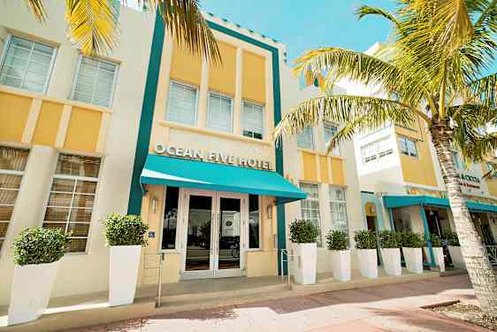 Hotellirakennuksen julkisivu, jossa on turkoosi markiisi ja keltaisia yksityiskohtia, ympäröity palmuilla. (Tekoälyn luoma teksti)