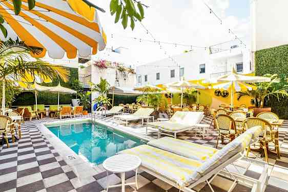 Valoisa ja värikäs ulkouima-allas, jota ympäröivät aurinkotuolit, aurinkovarjot ja istumapaikat vehreyden ja valkoisten rakennusten keskellä. (Tekoälyn luoma teksti)